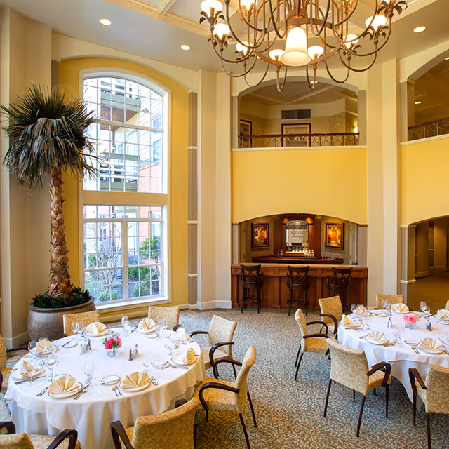 A bright and elegant dining room with round tables set with white tablecloths, folded napkins, glassware, and floral centerpieces. The room features large arched windows letting in natural light, a tall potted palm tree, a decorative chandelier, and a small bar area with three bar stools in the background.