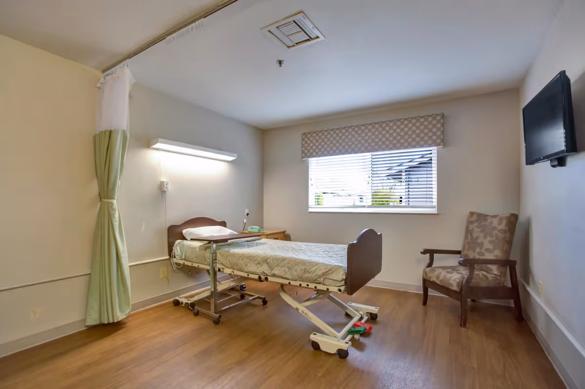 A simple patient room in a healthcare facility with a hospital bed, a small bedside table with a green phone, a patterned armchair, a wall-mounted TV, a window with blinds and a valance, and a green privacy curtain partially drawn to the side.