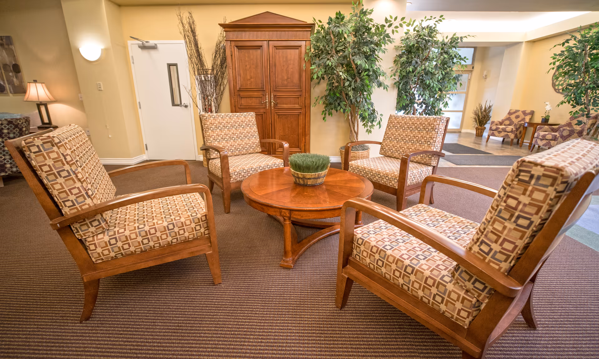 A cozy seating area in a senior living facility with four patterned armchairs arranged around a round wooden coffee table with a small green plant centerpiece. The room features beige walls, a wooden cabinet, potted plants, and additional seating in the background.