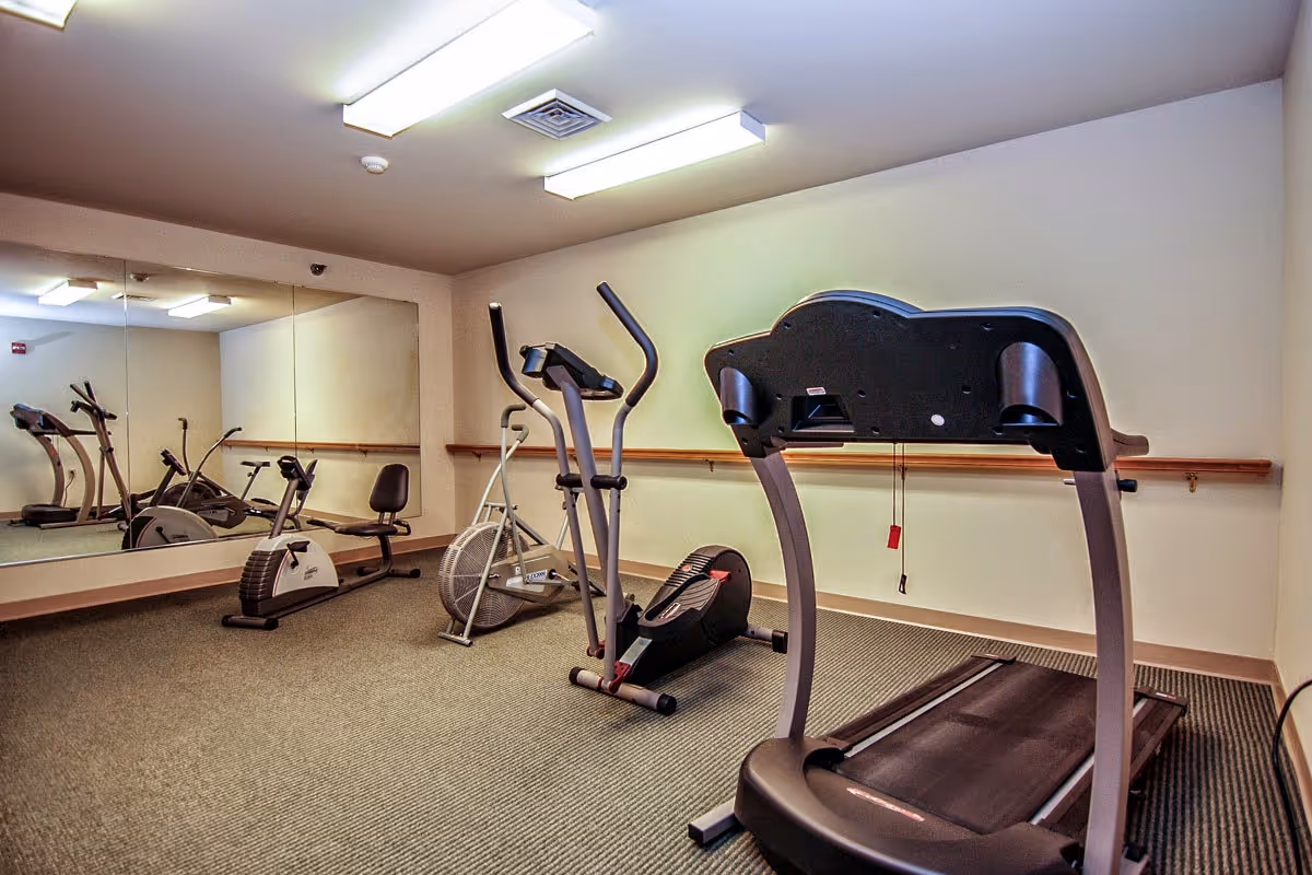 A small fitness room with exercise equipment including a treadmill, elliptical machine, stationary bike, and a recumbent bike. The room has beige walls, a large mirror on one side, carpeted floor, and fluorescent ceiling lights.
