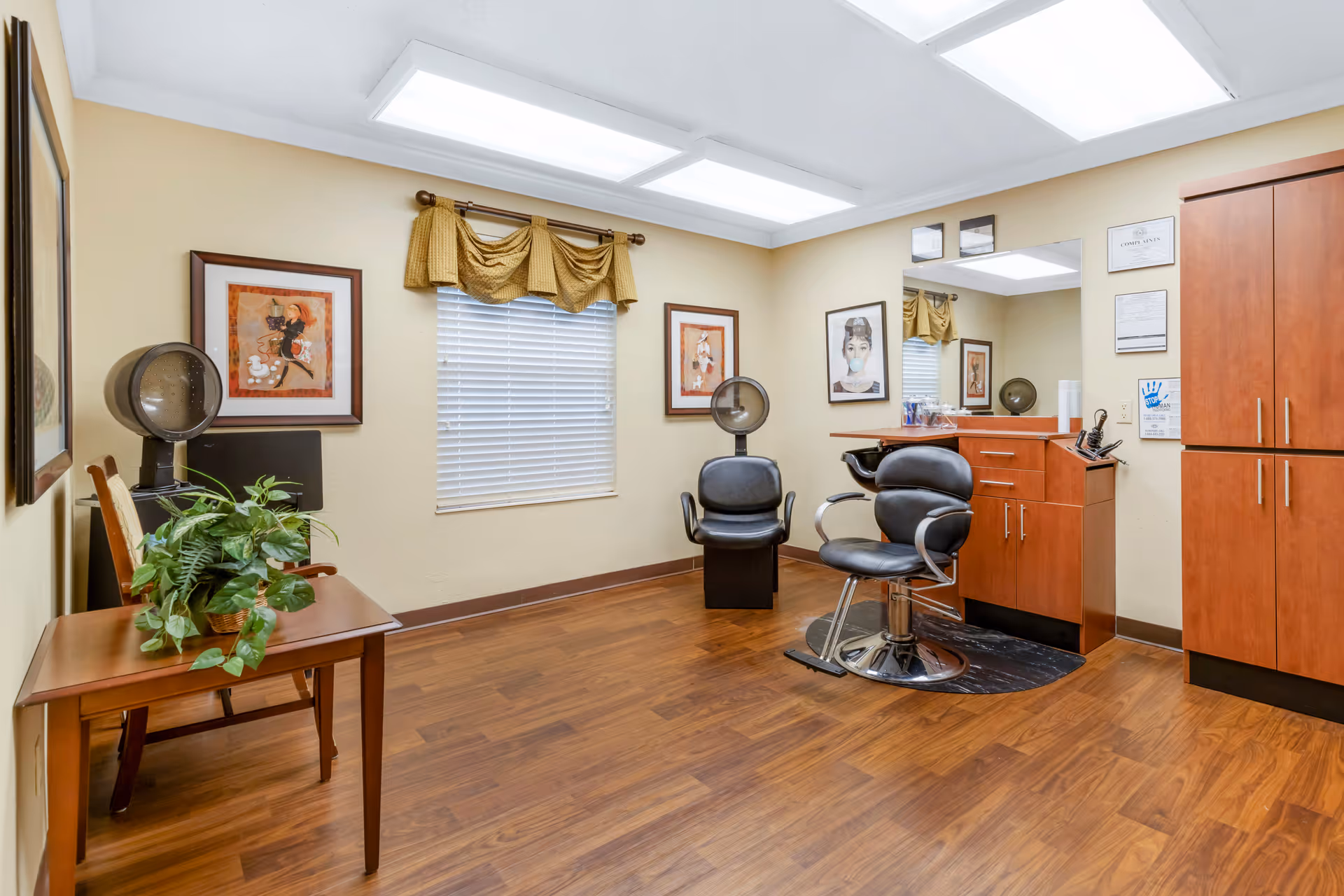 Small hair salon in a senior living facility with styling chairs, hood dryers, cabinetry, and framed artwork.