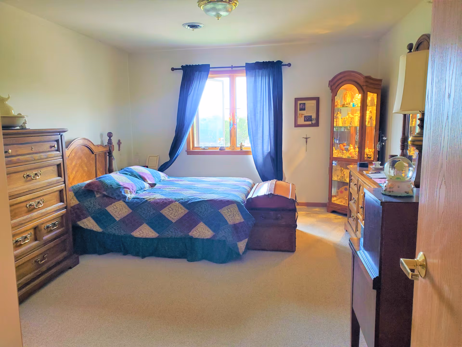 A cozy bedroom with a wooden bed frame and a colorful quilt on the bed. There is a wooden chest at the foot of the bed, a tall wooden dresser on the left, and a glass display cabinet filled with decorative items on the right. A window with dark curtains lets in natural light, and the walls are light-colored.