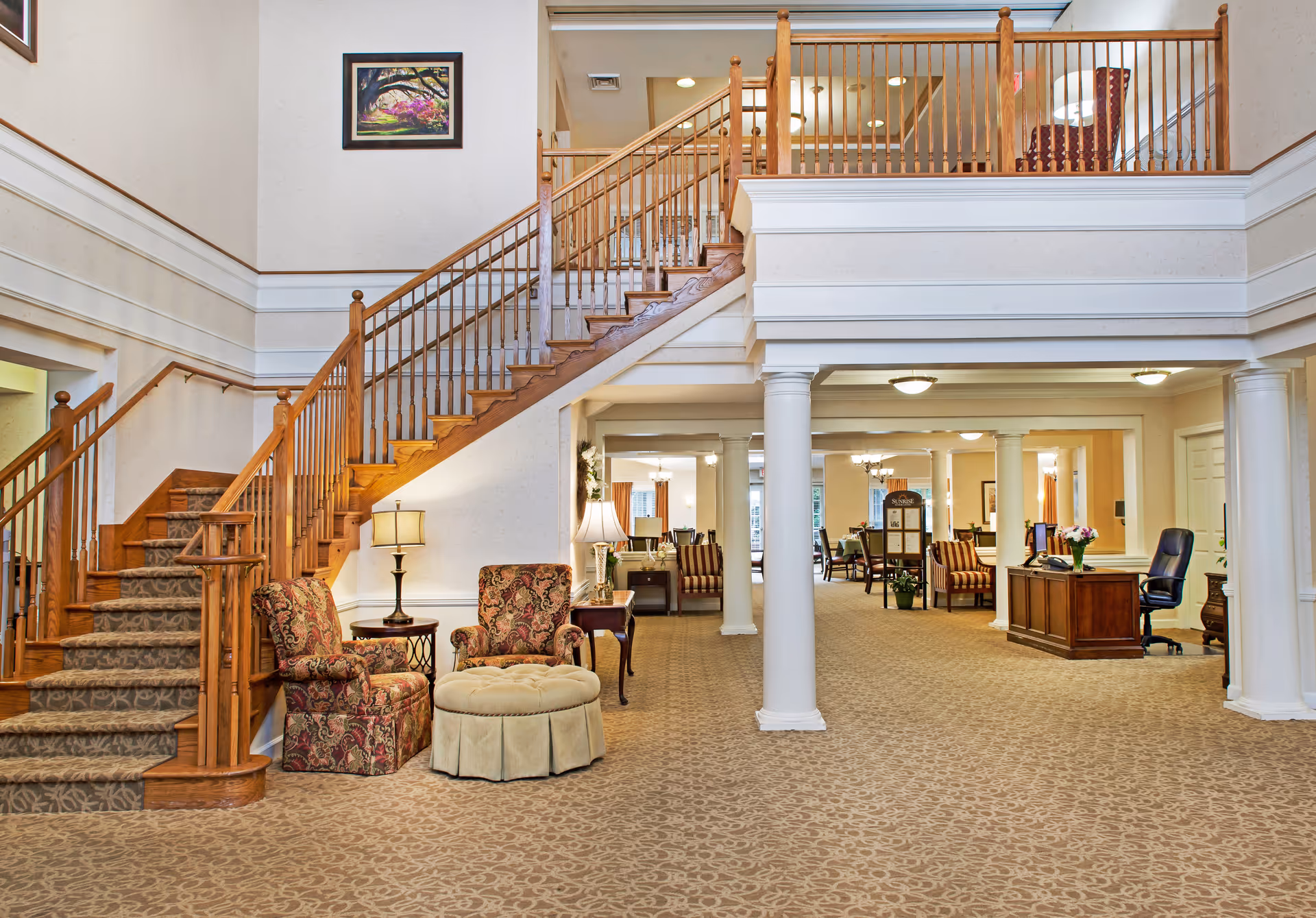 Open two-story lobby with a wooden staircase, upholstered chairs, columns, and a reception desk.