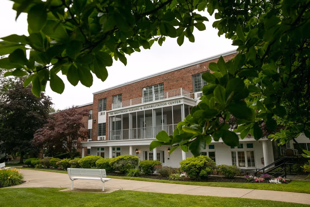 Exterior view of a brick building with white trim labeled The Brentwood Nursing & Rehabilitation Center, surrounded by green trees and bushes with a white bench on a paved walkway in the foreground.