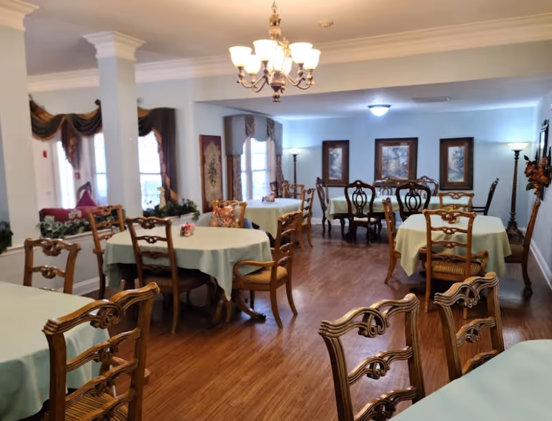 Dining room with round tables covered in light green tablecloths, wooden chairs, a central chandelier, and framed artwork on the walls.