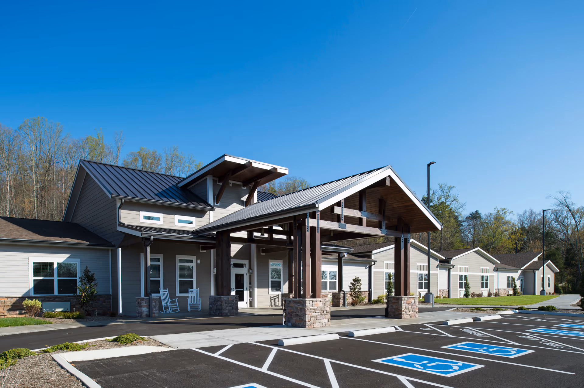 Exterior view of Pinnacle Assisted Living facility showing a modern single-story building with a covered entrance supported by wooden beams and stone pillars. The parking lot in front has multiple handicap parking spaces, and the sky is clear and blue.