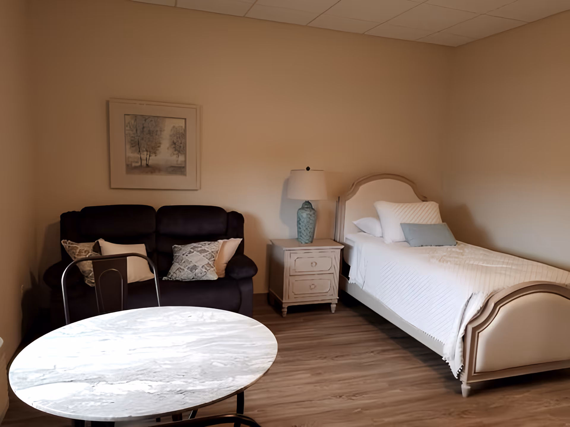 A furnished bedroom with a single bed, nightstand and lamp, a dark loveseat, and a round table.