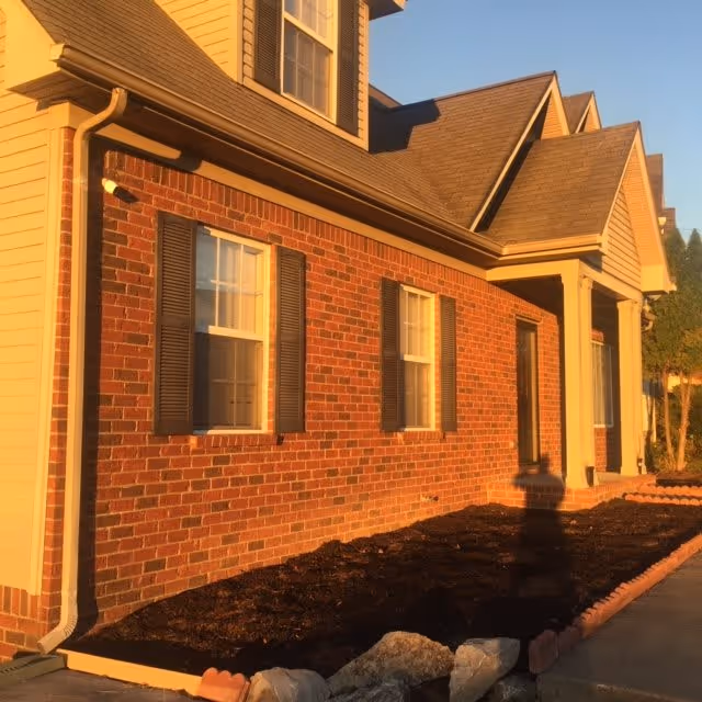 Brick-front residential building exterior with two windows, a covered front porch, and a mulched planting bed at sunset.
