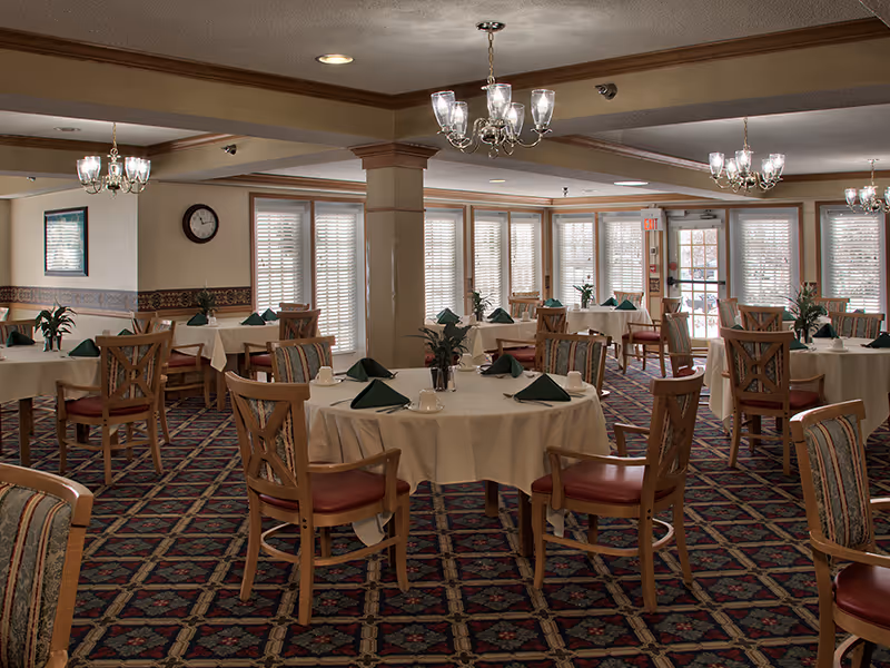 A dining room with round tables covered in white tablecloths, each set with green folded napkins, cups, and small plants as centerpieces. Wooden chairs with red cushions surround the tables. The room has patterned carpet flooring, multiple chandeliers hanging from the ceiling, and large windows with blinds letting in natural light.