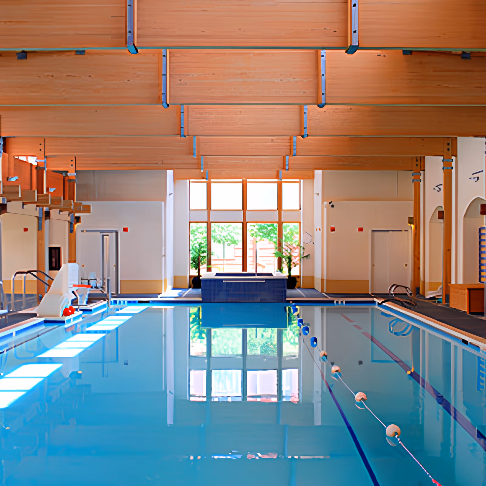 Indoor swimming pool with wooden-beamed ceiling, lane ropes, a pool lift, and a large window at the far end.