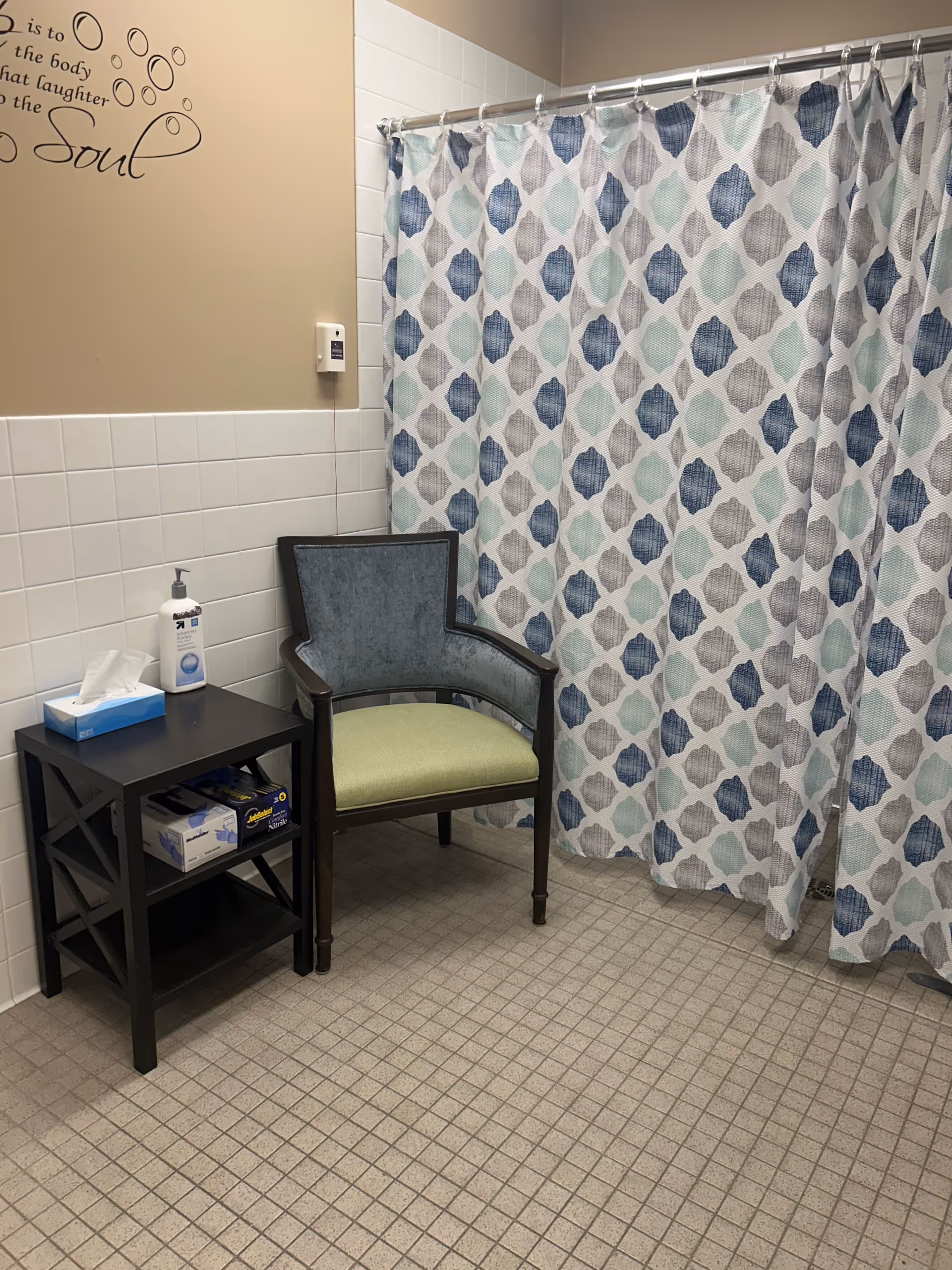 A bathroom corner with a patterned shower curtain in shades of blue, gray, and white. Next to the shower curtain is a wooden chair with a green seat cushion and blue backrest. A small black side table holds a box of tissues, a bottle of hand sanitizer, and a box of disposable gloves. The walls are tiled halfway up with white tiles and painted beige above. Part of a wall decal with a quote is visible.