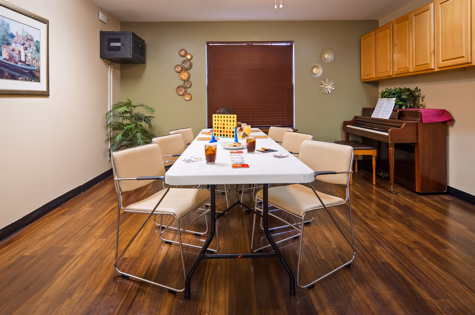 A room with a long white table surrounded by beige chairs on a wooden floor. On the table are drinks, playing cards, and a Connect Four game. The room has a piano with sheet music on the right side, a plant in the corner, wall decorations, and a window with closed blinds.