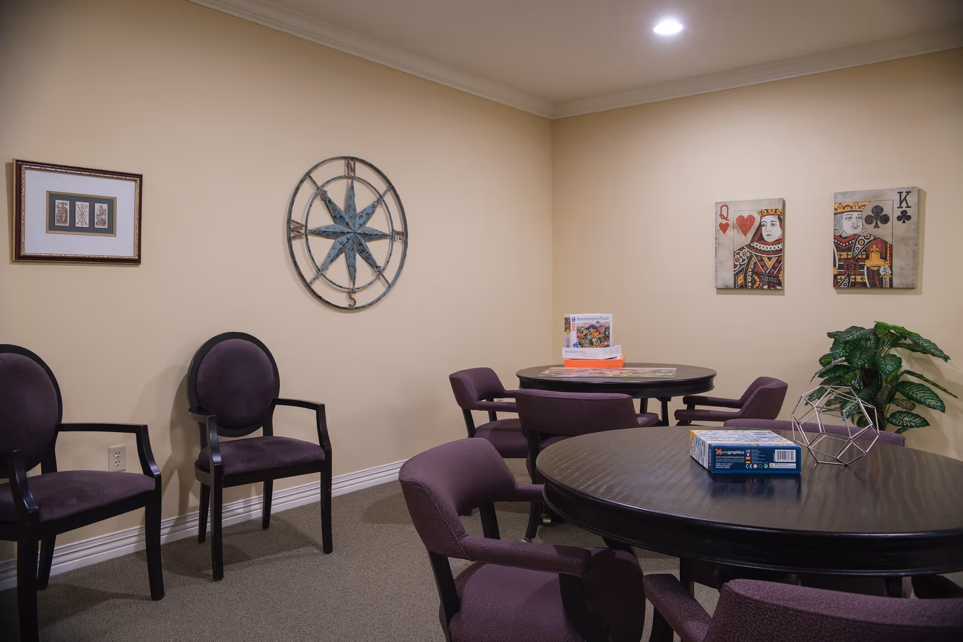 A cozy interior room with beige walls and carpeted floor, featuring two round dark wooden tables surrounded by purple upholstered chairs. On the tables are puzzle boxes and a geometric metal decor piece. The walls are decorated with framed artwork including a compass design and playing card images. A green potted plant is placed in the corner under a ceiling light.