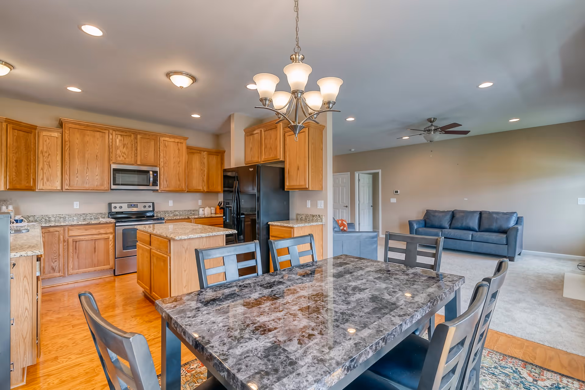 Open concept kitchen and living room area with wooden cabinets, stainless steel appliances, a black refrigerator, a marble dining table with six chairs, a chandelier above the table, and a blue sofa in the living room area.