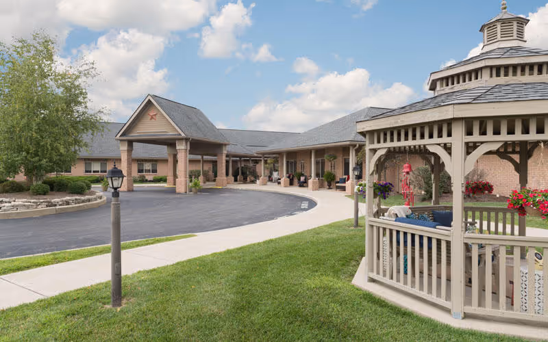 Exterior view of a single-story senior living facility with a covered entrance, a curved driveway, a well-maintained lawn, a lamp post, and a wooden gazebo with hanging flower baskets on the right side under a partly cloudy sky.