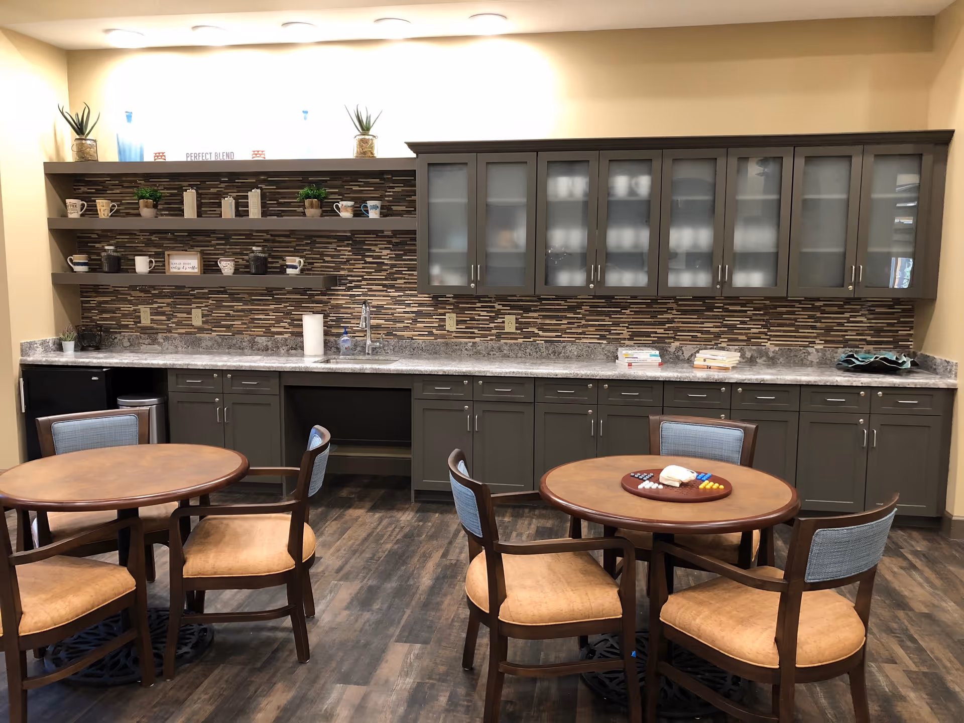 A cozy kitchen area with two round wooden tables each surrounded by four chairs with cushioned seats and backs. The back wall features a long countertop with a sink, gray cabinets below and above with frosted glass doors, and open shelves holding mugs, small plants, and decorative items. The backsplash has a mosaic tile design in shades of brown and beige. The floor is wood-patterned.