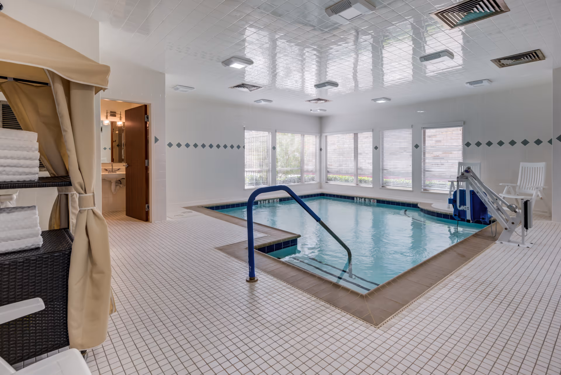 Indoor therapy-style swimming pool with a handrail, pool lift, windows, and stacked towels on shelving.