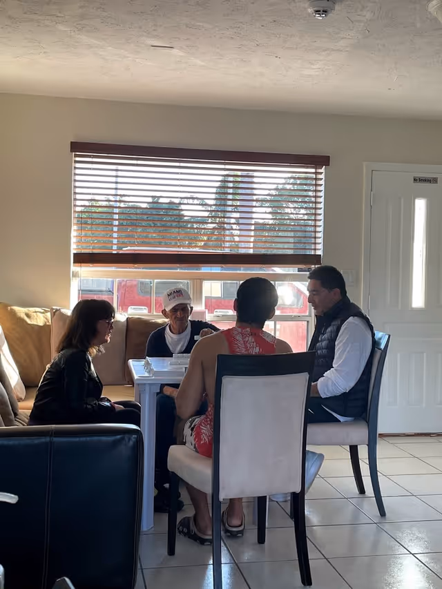 Four people sitting around a small white table in a room with a large window covered by horizontal blinds. Three men and one woman are engaged in an activity, possibly playing a game or having a discussion. The room has tiled floors, a beige couch, and a white door with a 'No Smoking' sign.