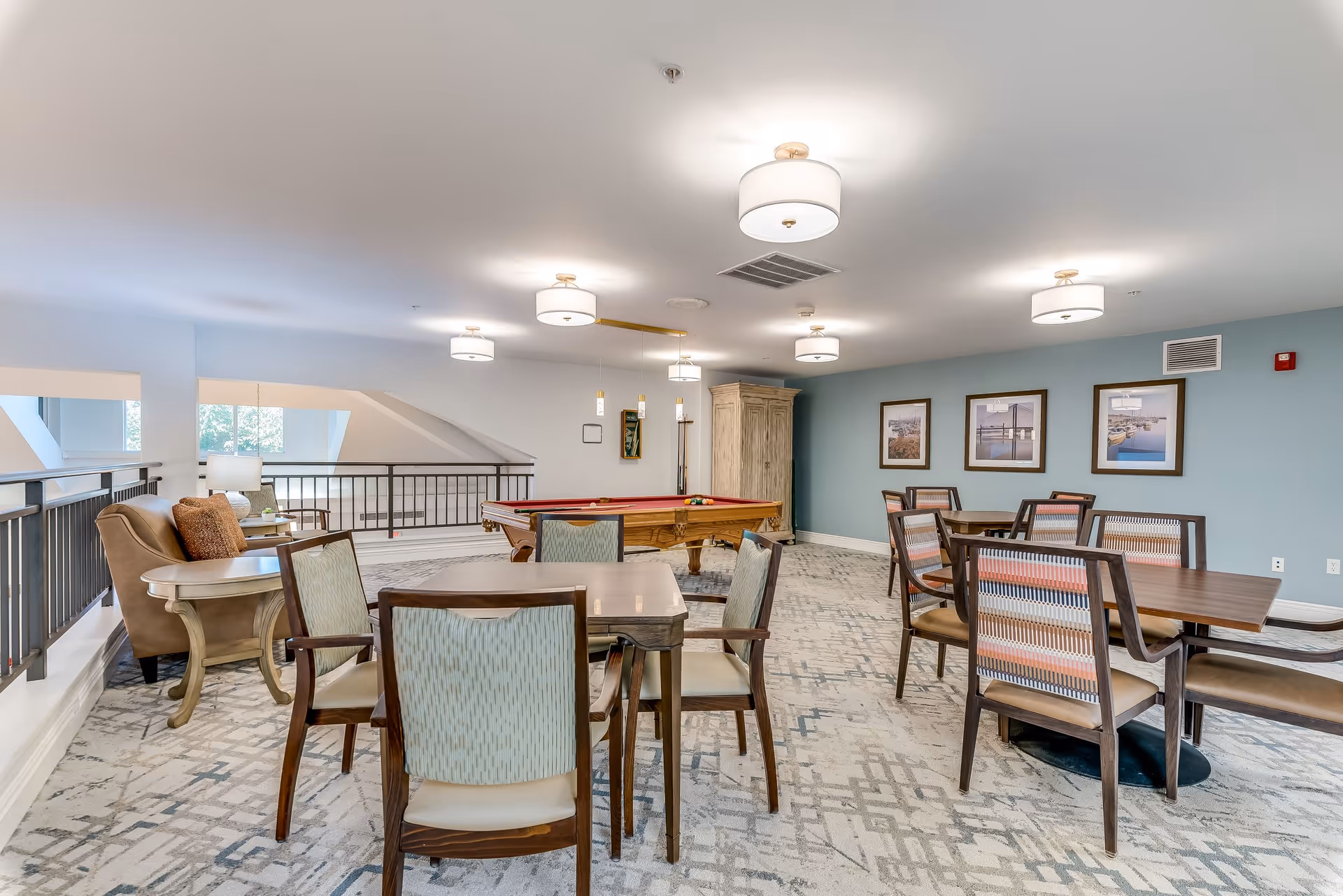 A spacious senior living common area with multiple tables and chairs arranged for socializing or activities. There is a pool table in the background, comfortable armchairs with side tables on the left, and framed pictures on a blue accent wall. The room is well-lit with ceiling lights and has a patterned carpet floor.