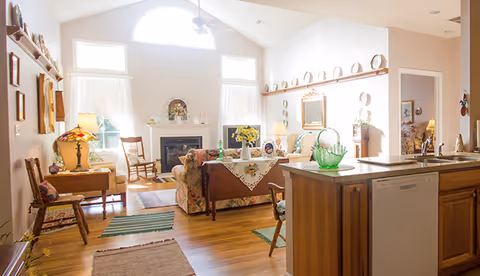 A bright and cozy living room area with wooden flooring, a fireplace with decorative items on the mantel, floral patterned sofas, a rocking chair, and a wooden side table with a lamp. The room has large windows with white curtains allowing natural light to fill the space. Adjacent to the living room is a kitchen area with wooden cabinets, a dishwasher, and a countertop with a green glass bowl and a vase of yellow flowers.