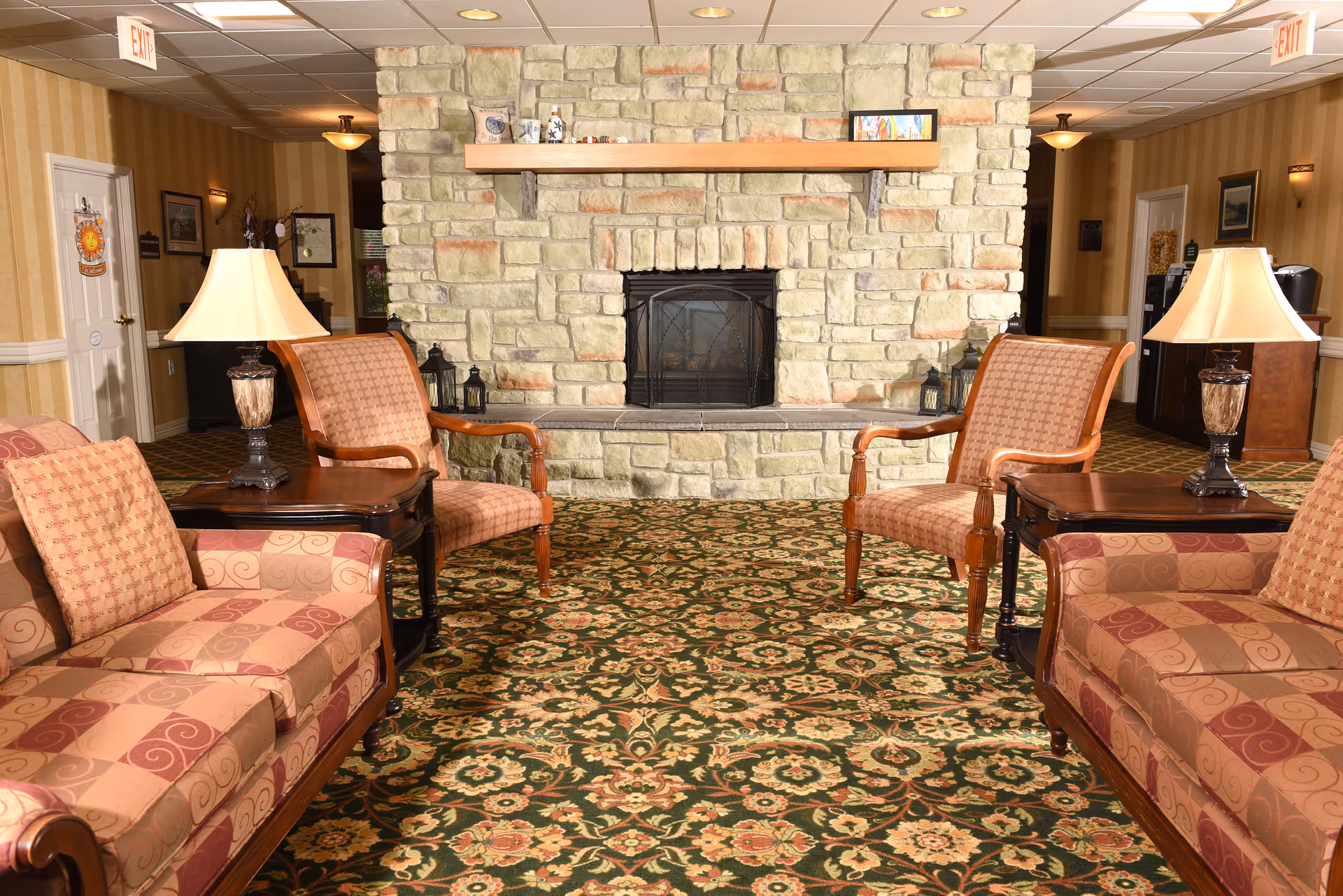 A cozy sitting area in a retirement community featuring two patterned sofas and two armchairs arranged around a stone fireplace. The room has warm lighting with table lamps on wooden side tables, floral patterned carpet, and beige striped wallpaper. There are exit signs visible on the ceiling.