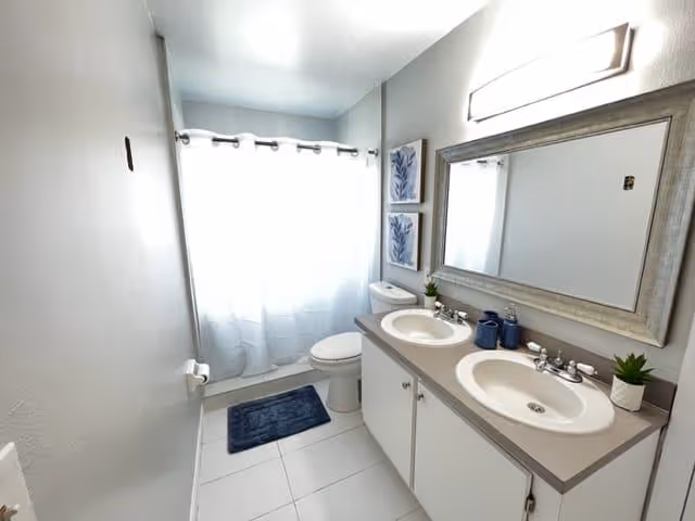 Bright bathroom with a double-sink vanity, large mirror, toilet, and a shower with a white curtain.