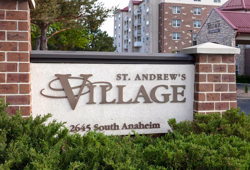 Stone entrance sign reading "St. Andrew's Village, 2645 South Anaheim" set among shrubs with the apartment building visible behind it.