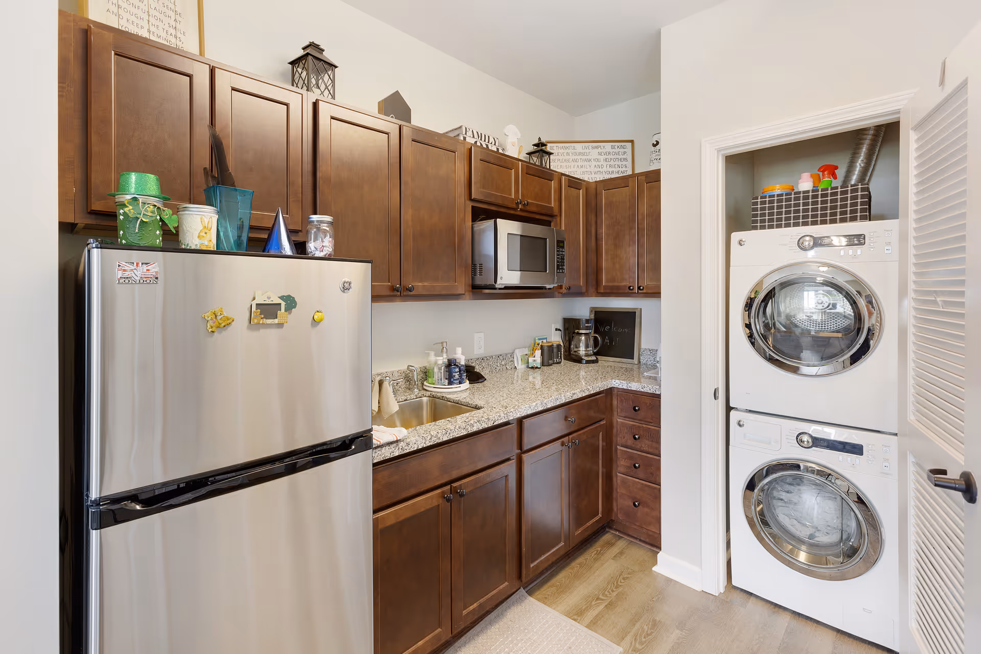 A compact kitchen area with dark wooden cabinets, a stainless steel refrigerator, a microwave mounted above the granite countertop, and a small sink. To the right, there is a closet with a stacked washer and dryer, along with cleaning supplies on top. The floor is light wood, and the walls are painted white.