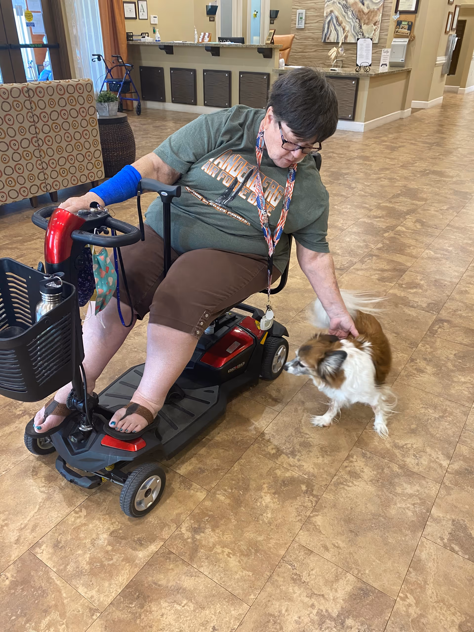 An elderly woman sitting on a mobility scooter inside a senior living facility. She is wearing glasses, a green t-shirt, brown shorts, and sandals. She is petting a small brown and white dog. The background shows a reception desk, chairs, and a walker near a window.