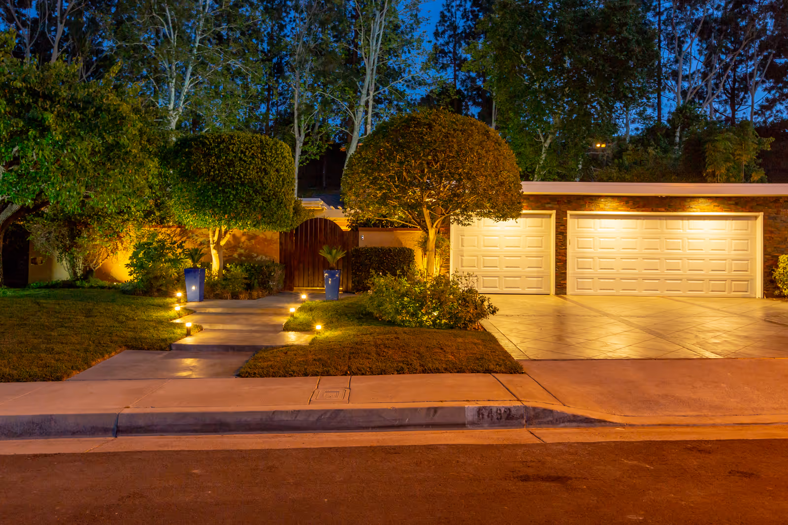 Evening view of a well-lit driveway and front yard of a house with neatly trimmed trees and bushes, a concrete pathway with small lights leading to a wooden gate, and a three-car garage with white doors.