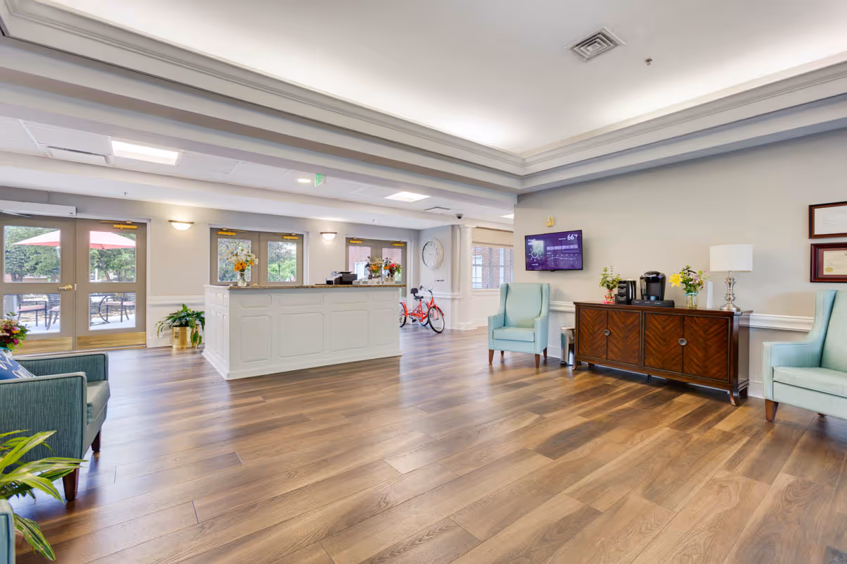 A spacious and bright reception area with wooden flooring, a white reception desk, light blue armchairs, a wooden cabinet with a coffee maker and flowers, a wall-mounted TV displaying weather information, and large windows and doors leading to an outdoor patio with tables and umbrellas.