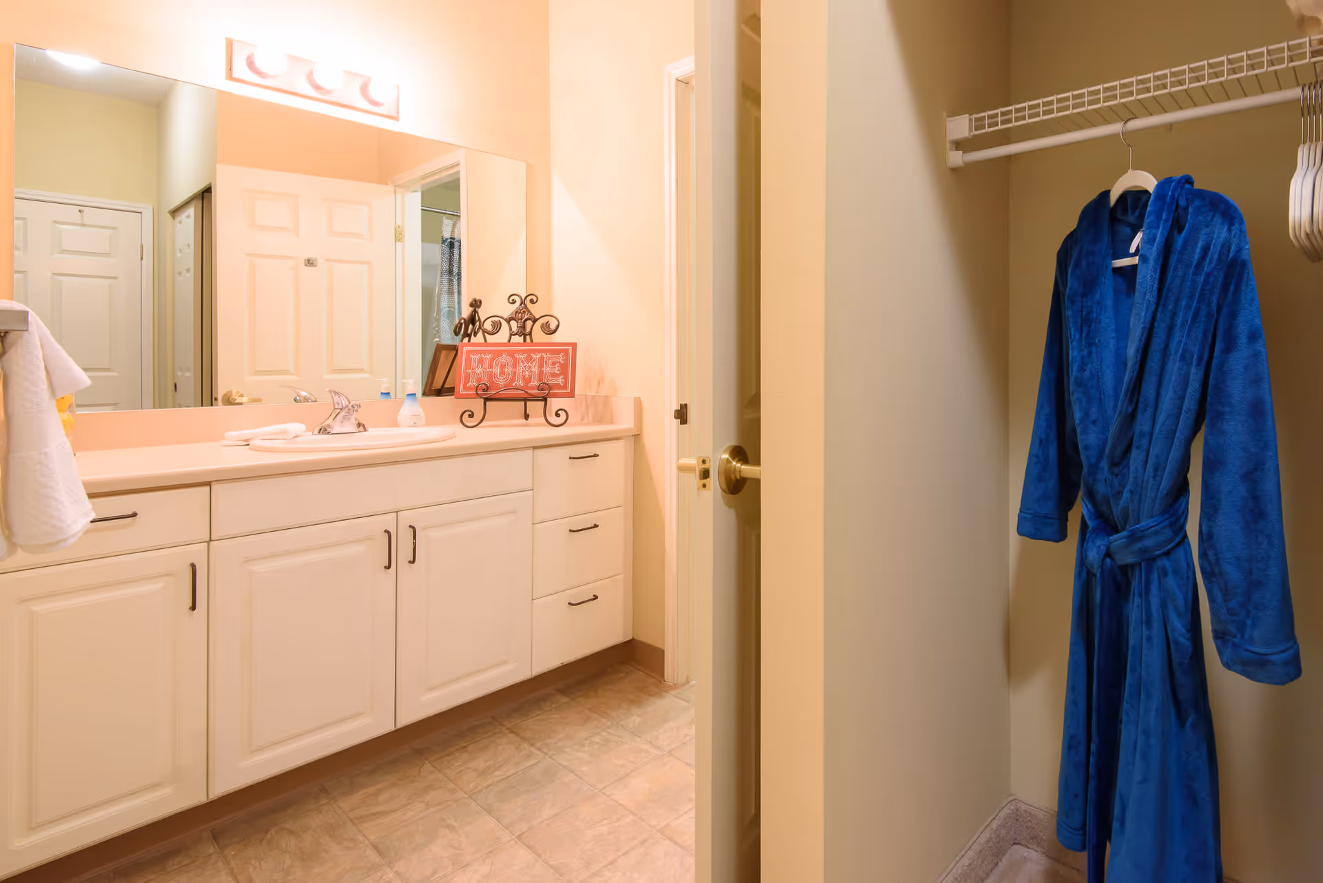A bathroom with a large mirror above a white vanity with multiple drawers and cabinets. On the countertop, there is a decorative sign that says 'HOME' and some toiletries. To the right, there is an open closet with a blue bathrobe hanging on a hanger. The floor is tiled and the walls are painted a light beige color.