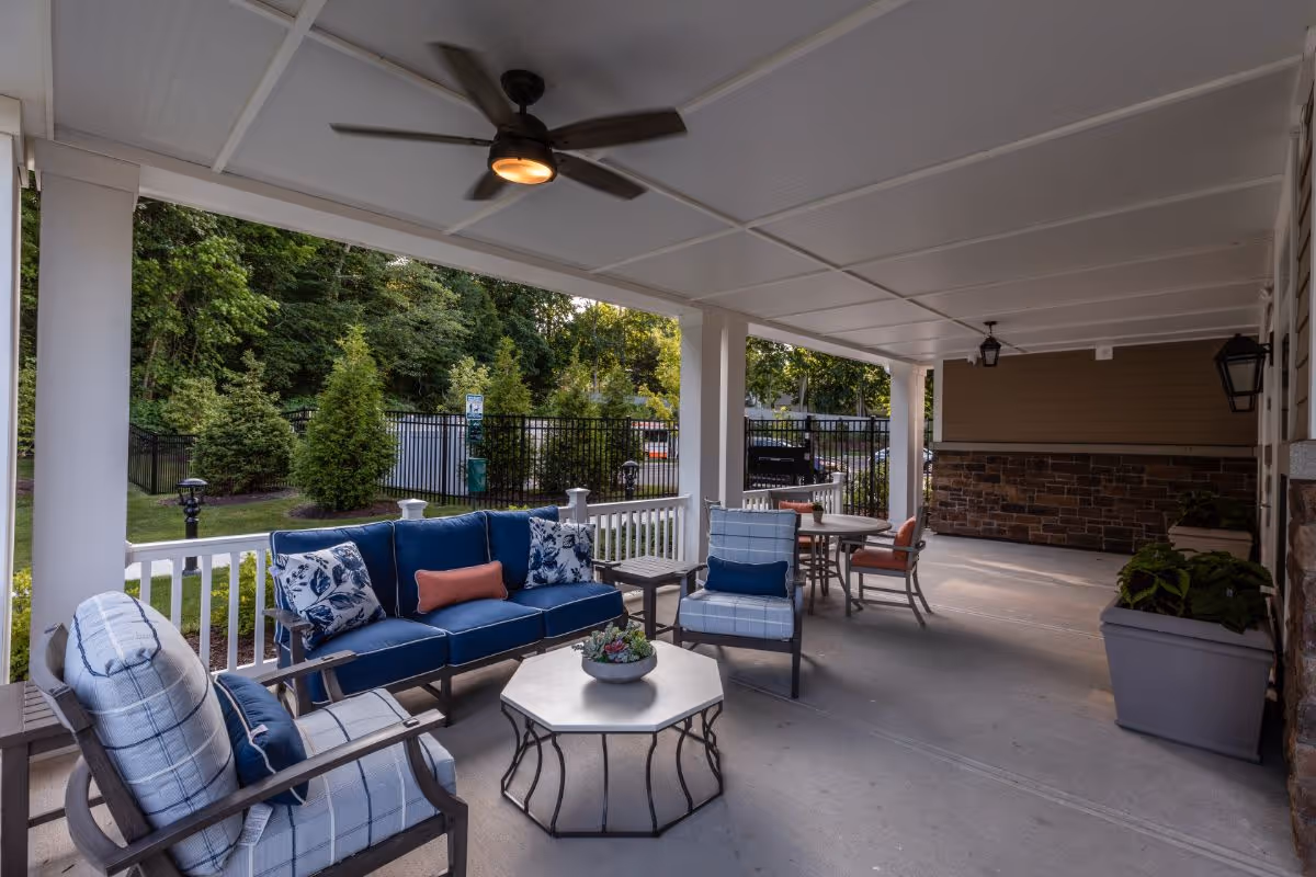 Covered outdoor patio area with cushioned seating including a blue sofa, armchairs with patterned cushions, a hexagonal coffee table, and a round dining table with chairs. The patio overlooks a fenced garden with trees and greenery.