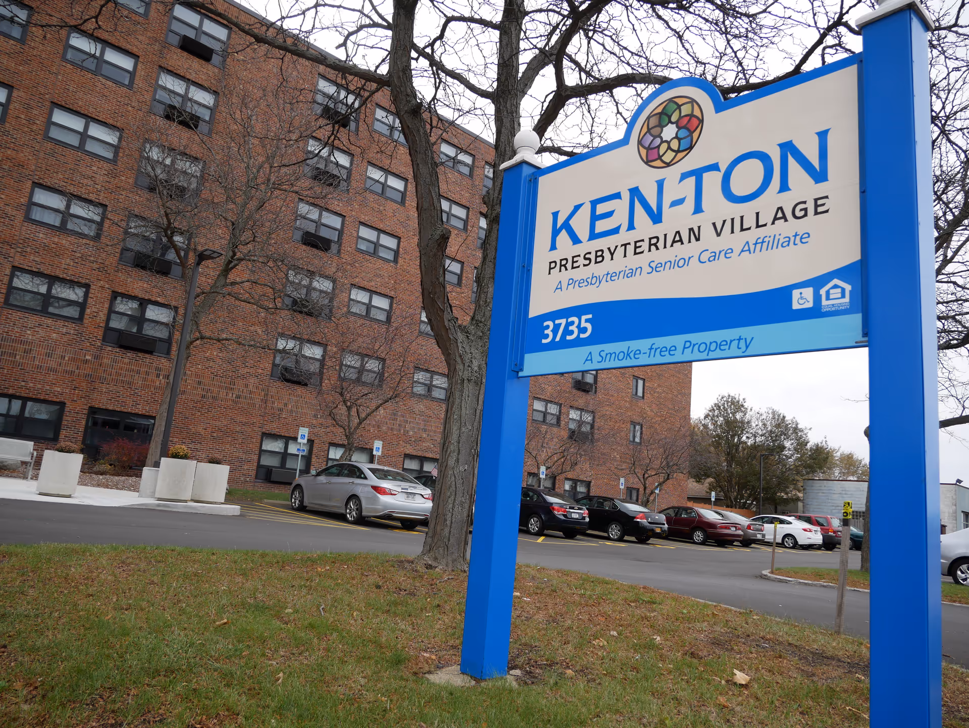 Outdoor view of Ken-Ton Presbyterian Village showing a large blue and white sign with the facility name and address, with a multi-story brick building and parked cars in the background.