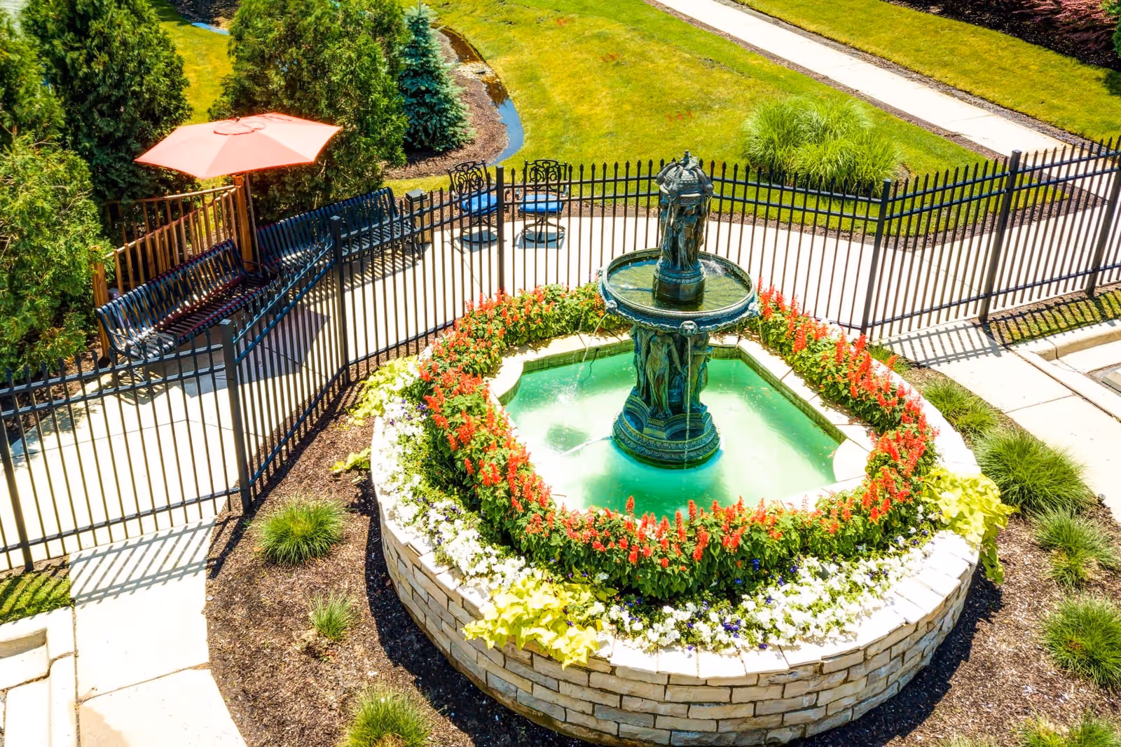 A circular stone fountain with water flowing from a central statue, surrounded by red and white flowers. The fountain is enclosed by a black metal fence. Nearby, there are benches, a red umbrella, and a paved walkway with green grass and shrubs around.