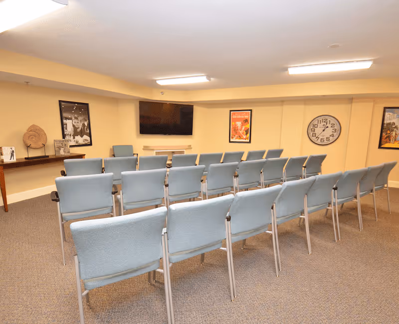 A small meeting or presentation room with rows of light blue chairs facing a wall-mounted flat screen TV. The room has beige walls, a carpeted floor, a large clock on the wall, and framed posters. There is also a wooden table with decorative items on the left side.