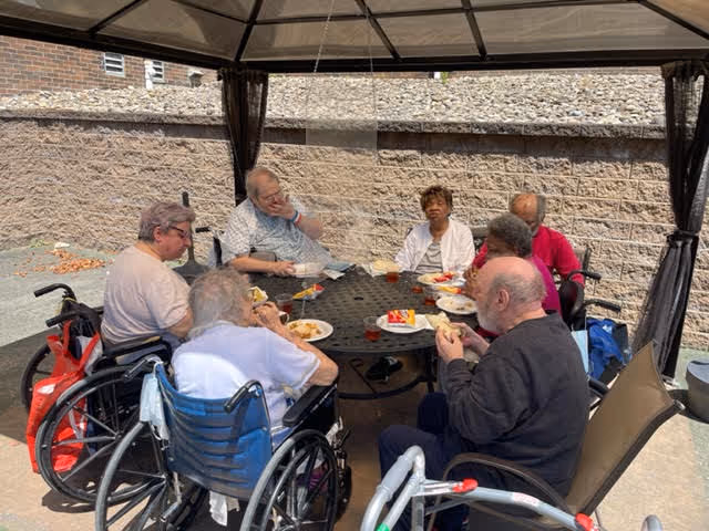 A group of elderly people, some in wheelchairs, sitting around a round outdoor table under a gazebo, eating and socializing. The setting is an outdoor patio area with a brick wall in the background.