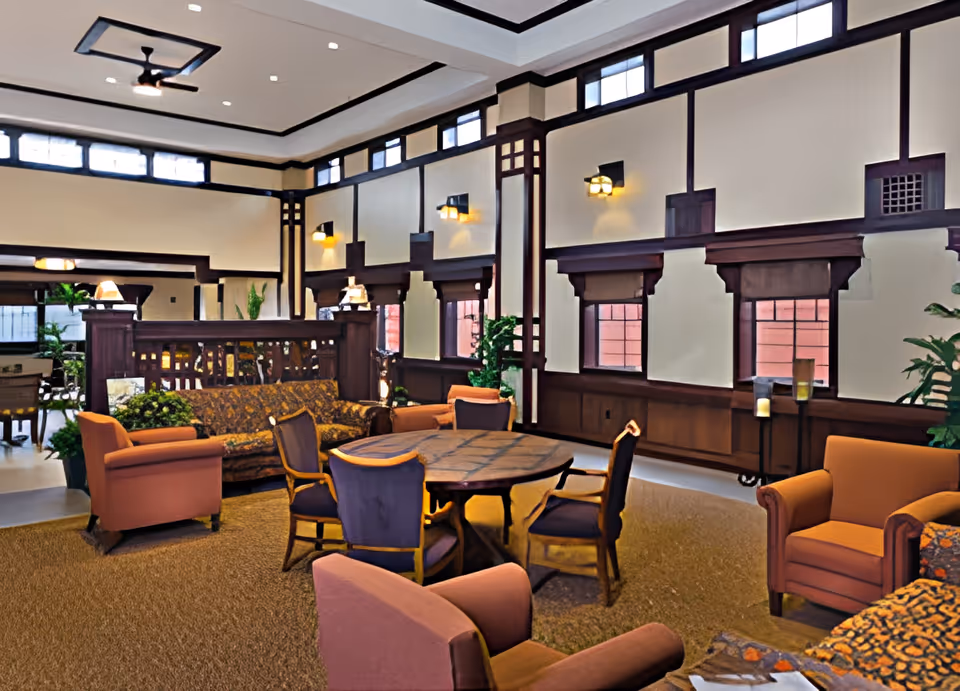 A spacious and well-lit living room area in Village Assisted Living featuring a round wooden table surrounded by four chairs, several upholstered armchairs, and a patterned sofa. The room has high ceilings with decorative wooden trim, multiple windows with shades, wall sconces providing warm lighting, and several potted plants adding greenery.