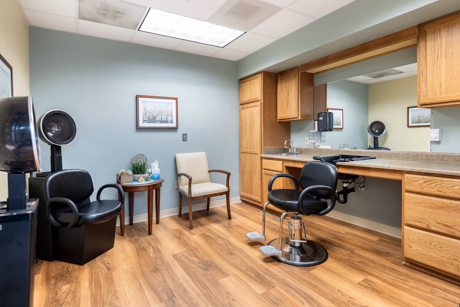 Interior view of a salon area in a senior living facility with two black salon chairs, a beige guest chair, a small round wooden table with decorative items, wooden cabinets, a countertop with a sink, and a large mirror on the wall.