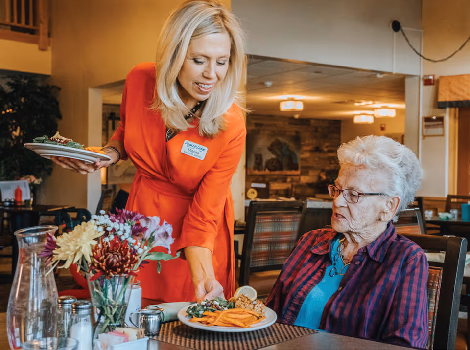 A woman in a red dress serving a plate of food to an elderly woman seated at a dining table in a warmly lit dining room. The table has a vase with colorful flowers and a glass pitcher of water.