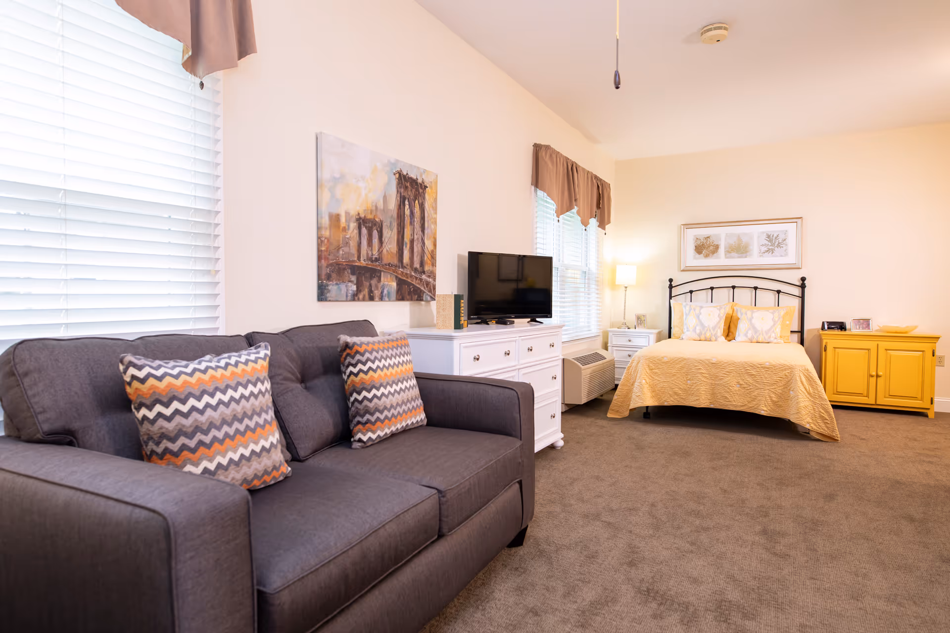A well-lit room featuring a gray sofa with two patterned pillows, a white dresser with a TV on top, a bed with a yellow bedspread and decorative pillows, a yellow cabinet, and framed artwork on the walls. The room has beige walls, carpeted floor, and windows with blinds and valances.