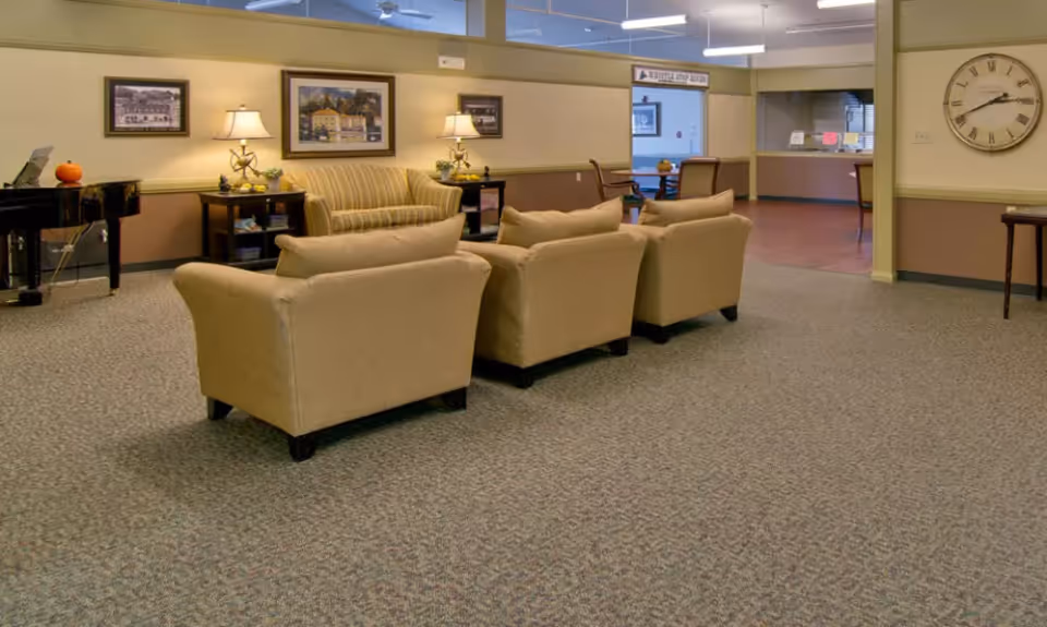 A cozy living room area in a senior living facility featuring a striped sofa and two beige armchairs arranged around a small table. The room has carpeted flooring, framed artwork on the walls, two table lamps on side tables, a large wall clock, and a black piano with a small pumpkin on it. In the background, there is an open doorway leading to another room with tables and chairs.