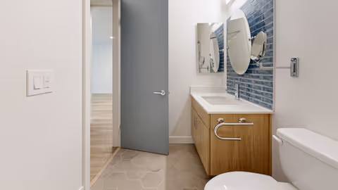 Modern bathroom with a wooden vanity featuring a white sink and a round mirror above it. The wall behind the sink is decorated with blue rectangular tiles. There is a toilet to the right and a partially open gray door leading to a room with wooden flooring.