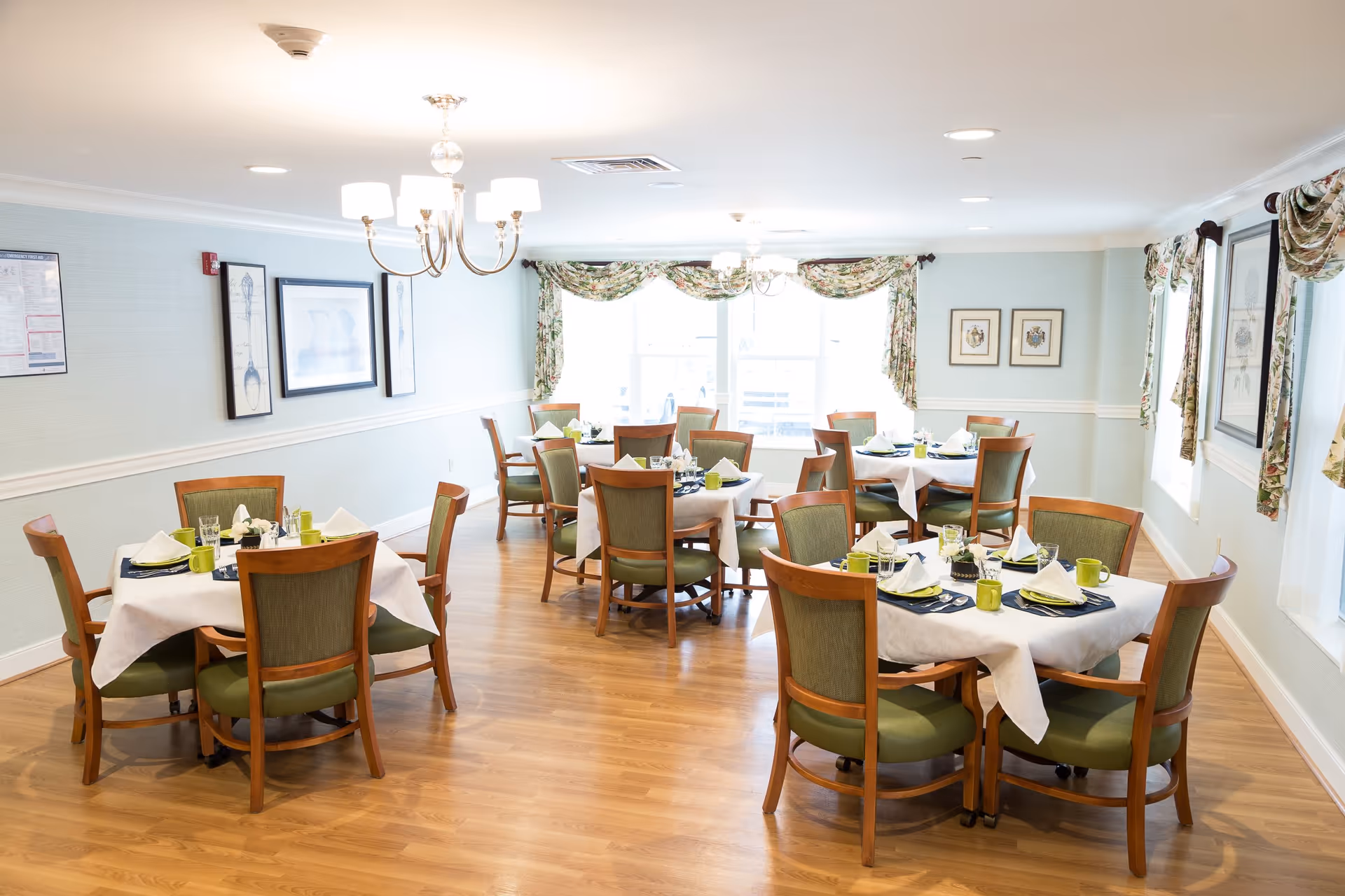 A bright dining room with several round tables covered with white tablecloths, each set with green mugs, napkins, and utensils. The room has wooden floors, green cushioned chairs, floral curtains on the windows, and framed artwork on the walls. A chandelier hangs from the ceiling providing light.