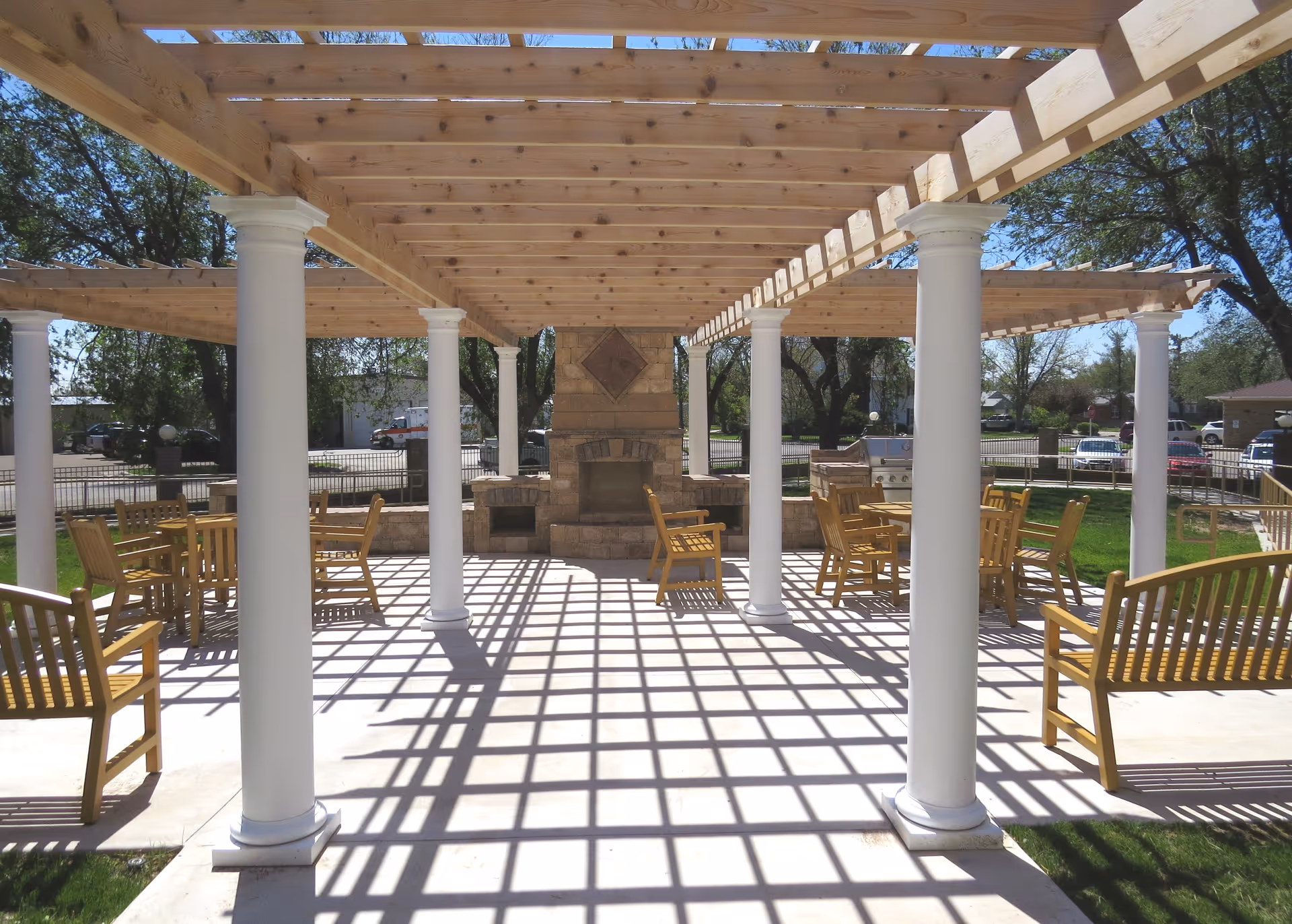 Outdoor patio area with wooden pergola supported by white columns, wooden benches and tables with chairs, and a stone fireplace in the background. Trees and parked cars are visible beyond the fenced area.