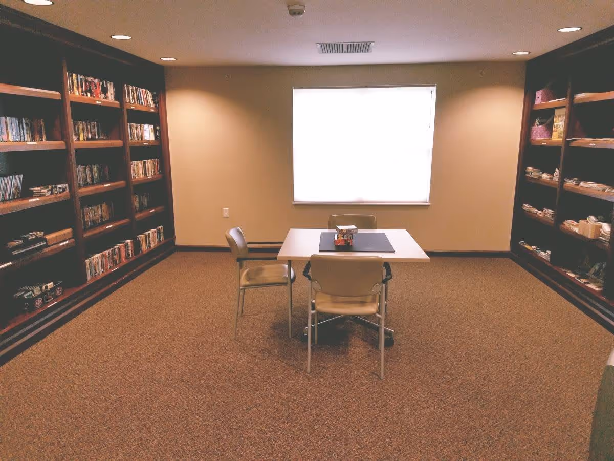 A quiet room with carpeted floor featuring a small square table with three chairs around it. The room has built-in wooden bookshelves on both sides filled with books and other items. A window with a white blind is centered on the far wall, allowing natural light to enter the room.