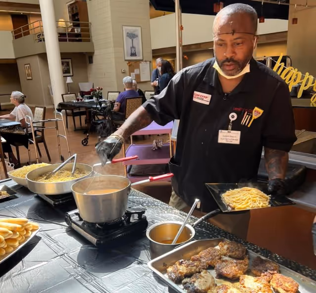 A staff member serves pasta and sauce at a buffet-style station in a communal dining area with seated residents in the background.