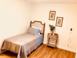 A simple bedroom with a single bed covered in a beige quilt and a matching pillow. Next to the bed is a small wooden nightstand with a potted plant on top. Two framed botanical prints hang on the wall above the nightstand. The floor is wooden and the walls are painted white.