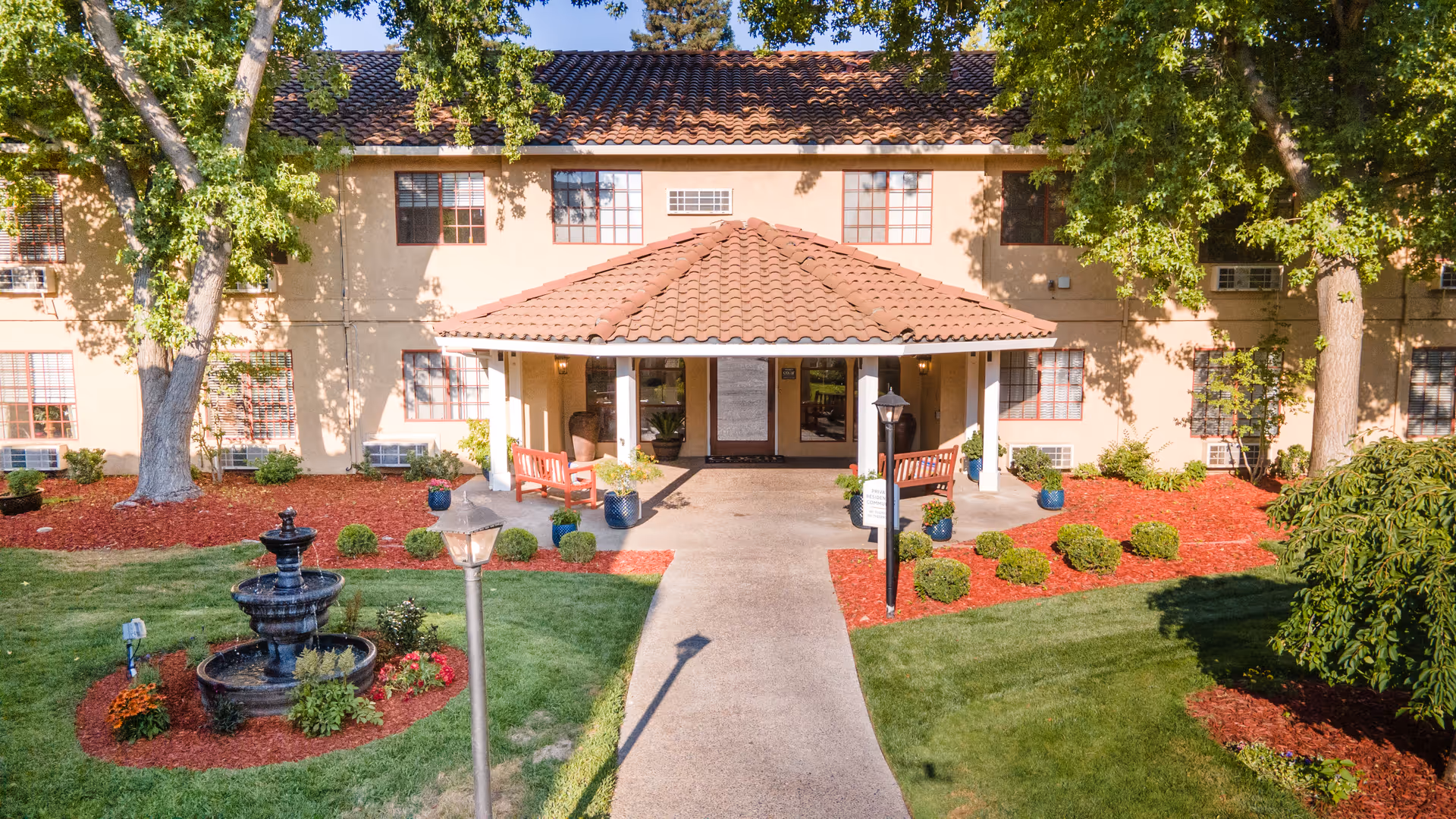 Front entrance of a two-story residential building with a tile-roofed portico, walkway, fountain, benches, and landscaped lawns.