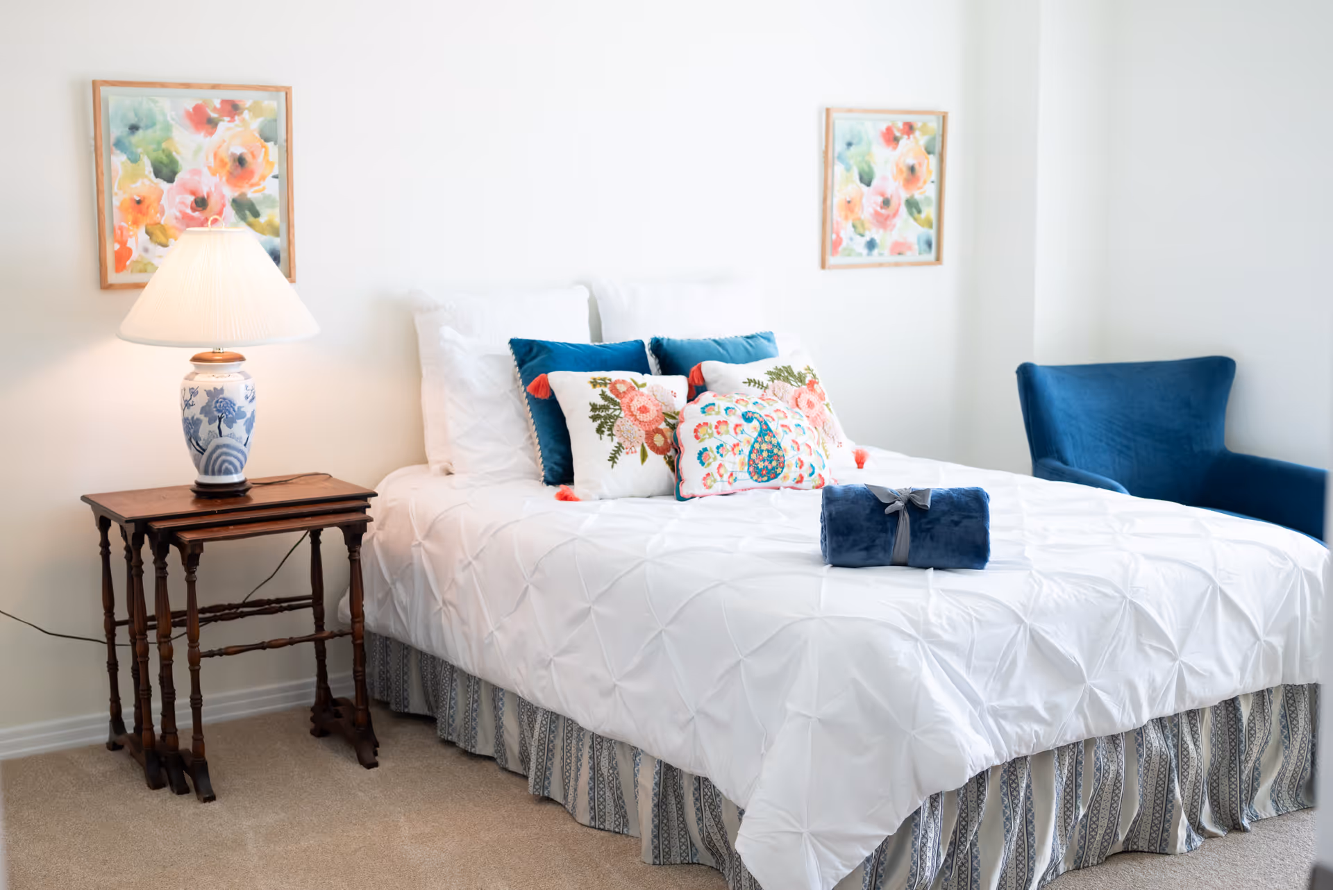 A bright bedroom with a neatly made bed featuring white bedding and decorative pillows with floral and peacock designs. There is a rolled blue blanket on the bed. On the left side, there is a wooden side table with a blue and white lamp. Two colorful floral paintings hang on the white walls above the bed. A blue upholstered chair is positioned in the corner on the right side.